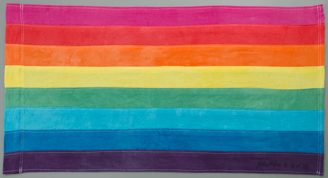 A hand-dyed fabric rainbow flag with eight horizontal stripes — hot pink, red, orange, yellow, green, turquoise, blue, and violet — photographed flat on a gray background, signed 'Gilbert Baker #3 of 35' in the bottom corner.