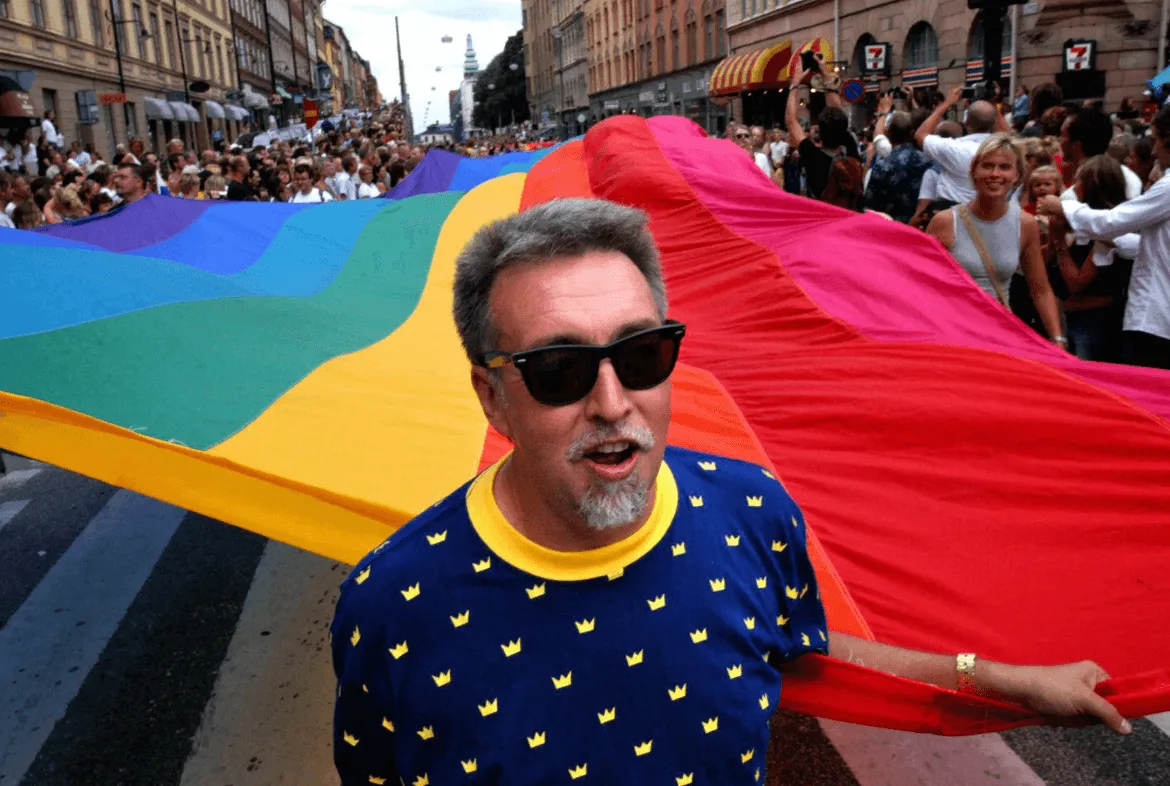 Gilbert Baker in sunglasses and a blue crown-patterned shirt at the head of a parade, an enormous rainbow flag held aloft by crowds filling the street behind him.