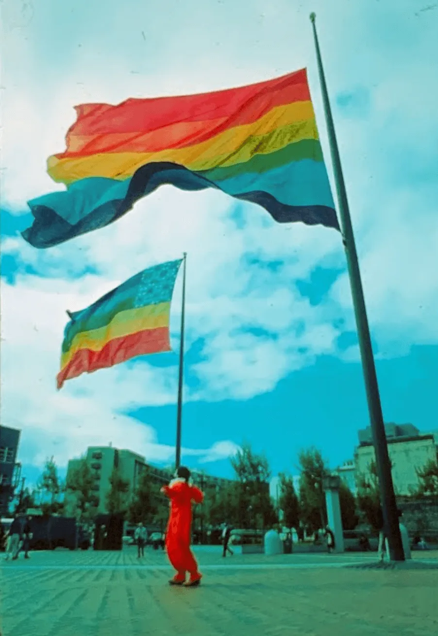 Two enormous eight-color rainbow flags flying from the flagpoles at San Francisco's United Nations Plaza in 1978. One flag carries only the eight horizontal rainbow stripes; the other is a stars-and-stripes variant with a tie-dyed star canton in its upper corner.