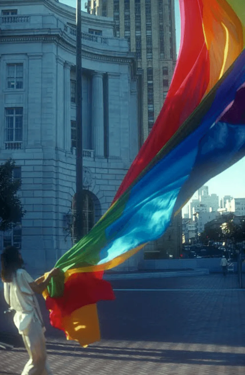 A person holds one of the original eight-stripe 1978 rainbow flags aloft in the wind at San Francisco's Civic Center, a classical columned building rising behind them.