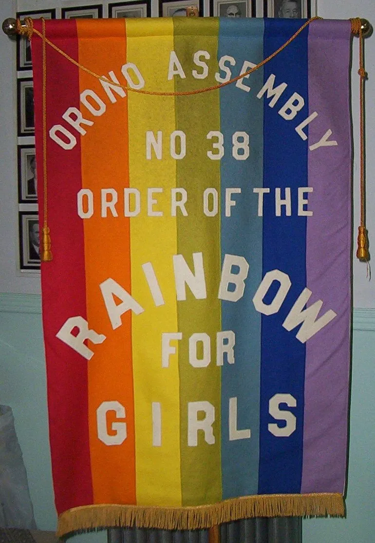 A vintage felt assembly banner of Orono Assembly No. 38, Order of the Rainbow for Girls, with seven vertical stripes — red, orange, yellow, green, light blue, dark blue, violet — gold cord and fringe, hung in a lodge room.