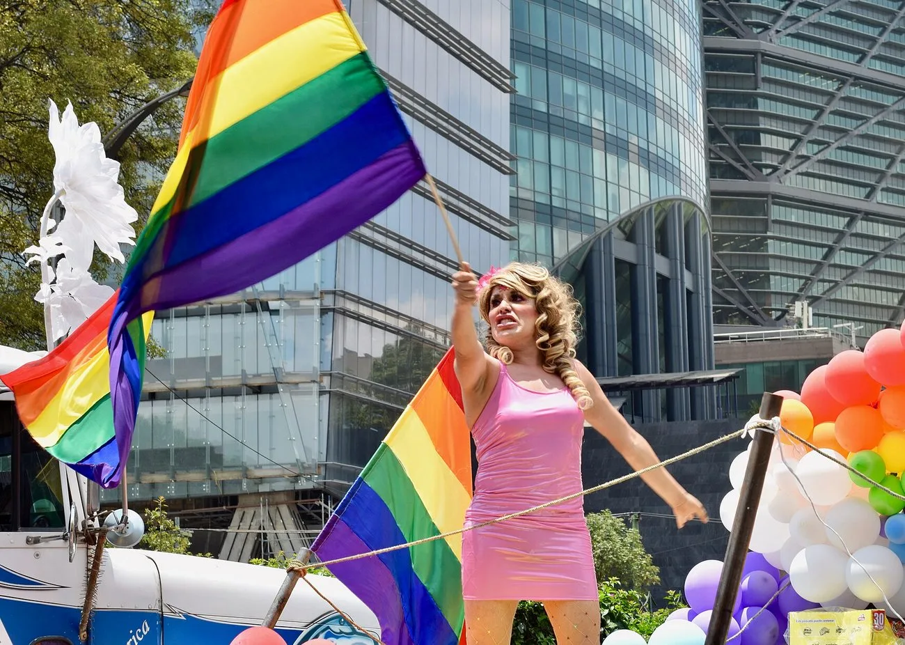 A performer in a pink dress and blonde wig waves a large rainbow flag from a parade float in front of a glass office tower, with balloon arches and a second rainbow flag beside her.