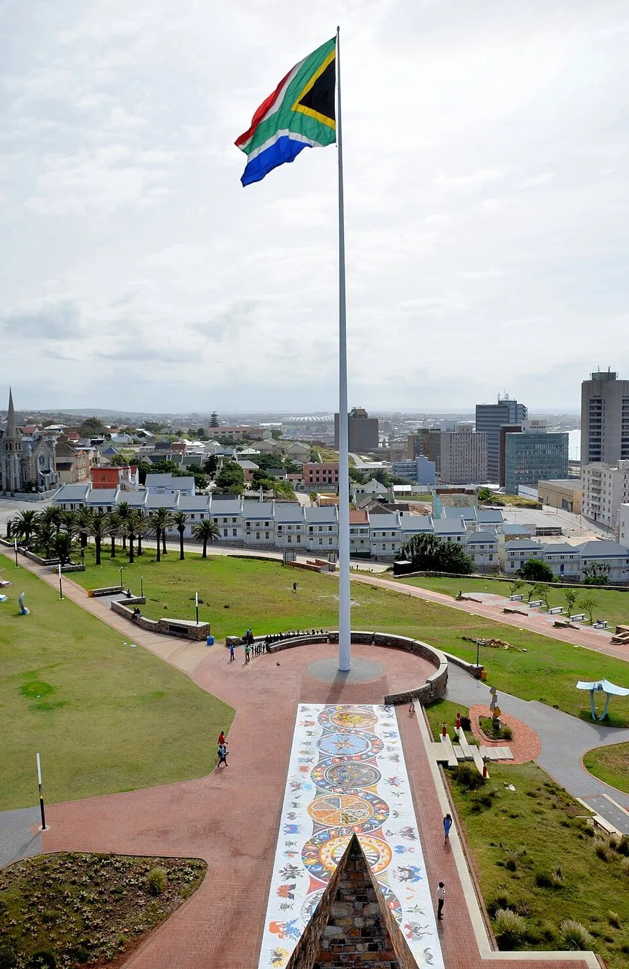 A very tall slender flagpole on a grassy hilltop reserve flying the South African national flag, seen from below against a clear sky.