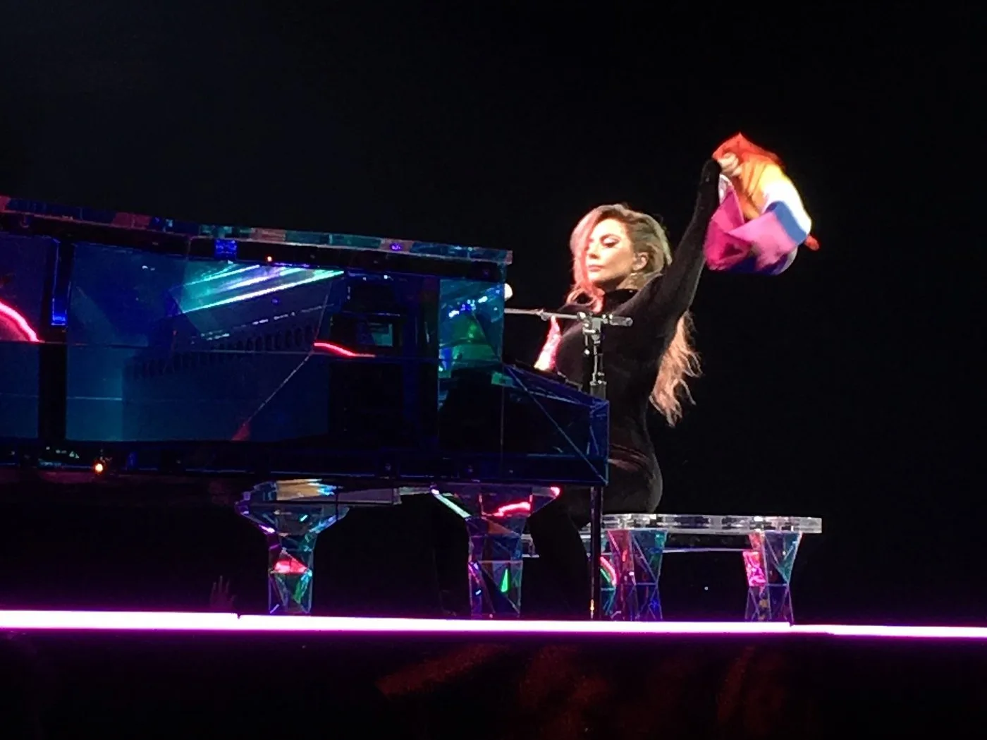 Lady Gaga seated at a mirrored grand piano on a darkened arena stage, raising a bunched rainbow flag overhead in one gloved hand.