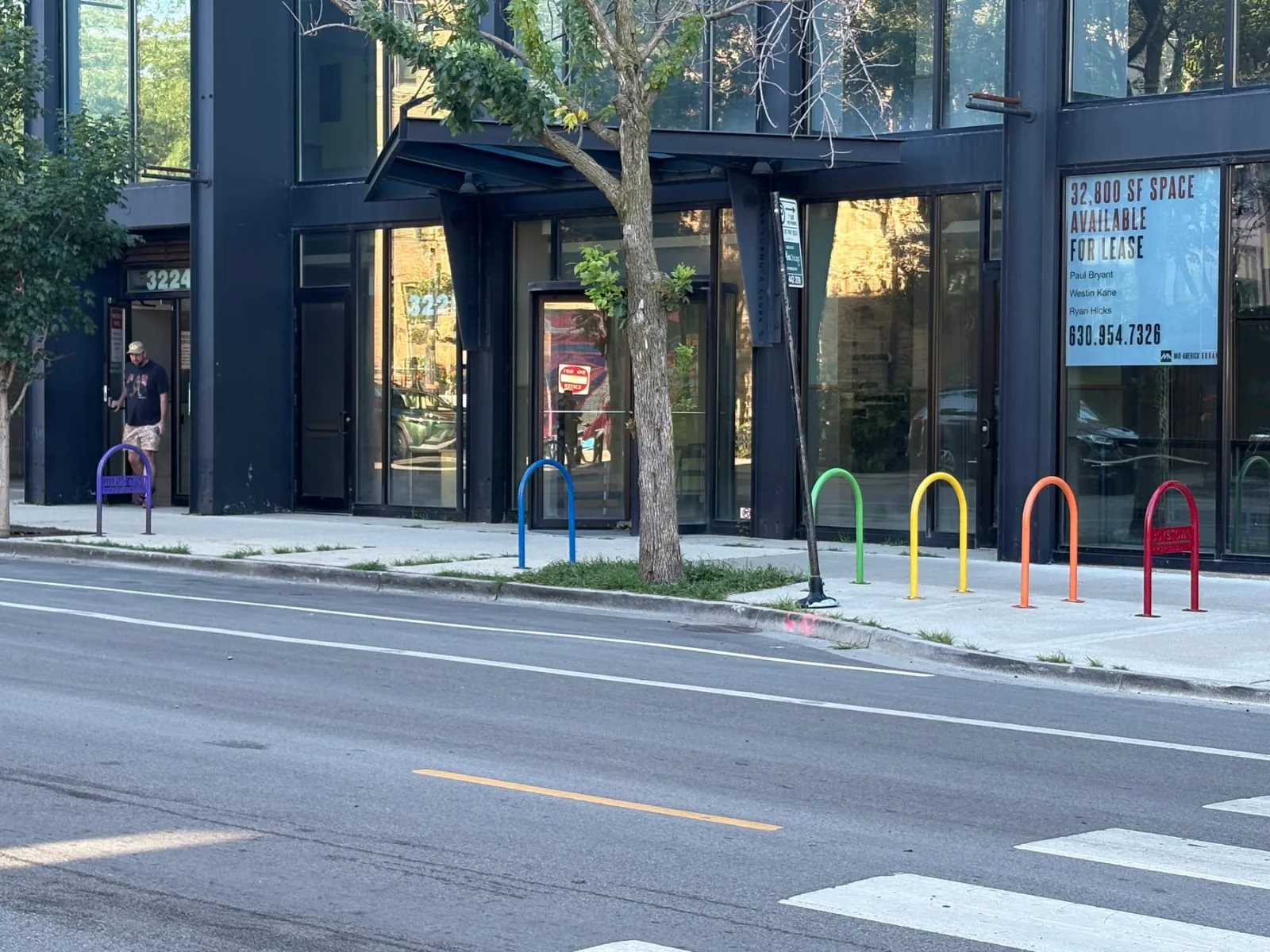 Six inverted-U bike racks on a Chicago sidewalk, each painted a different color of the six-stripe rainbow: violet, blue, green, yellow, orange and red, in front of a dark glass storefront numbered 3224.