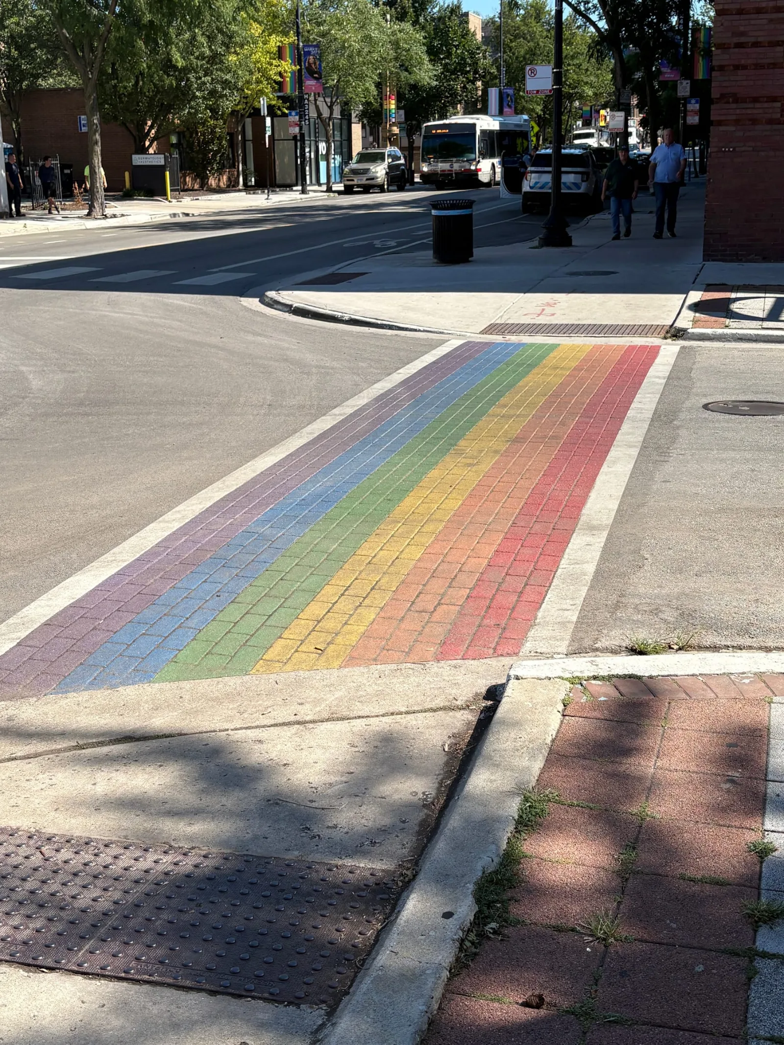 A six-stripe rainbow crosswalk painted across brick paving on North Halsted Street in Chicago, red nearest the far curb through violet, with a CTA bus and rainbow-topped pylons up the street behind it.