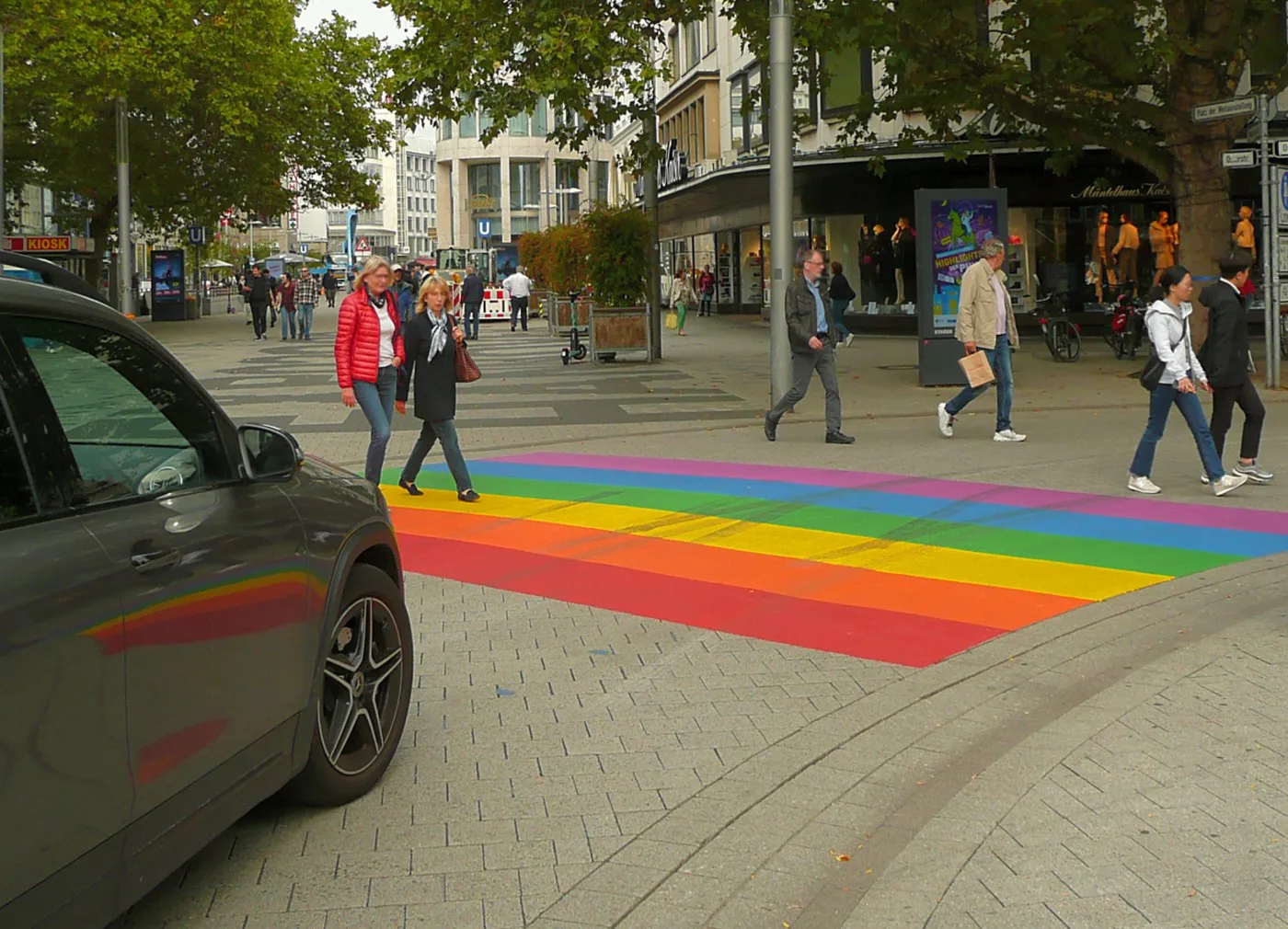 Pedestrians walking across a rainbow-striped crosswalk on Karmarschstraße in Hannover, Germany, with shops and trees lining the street.