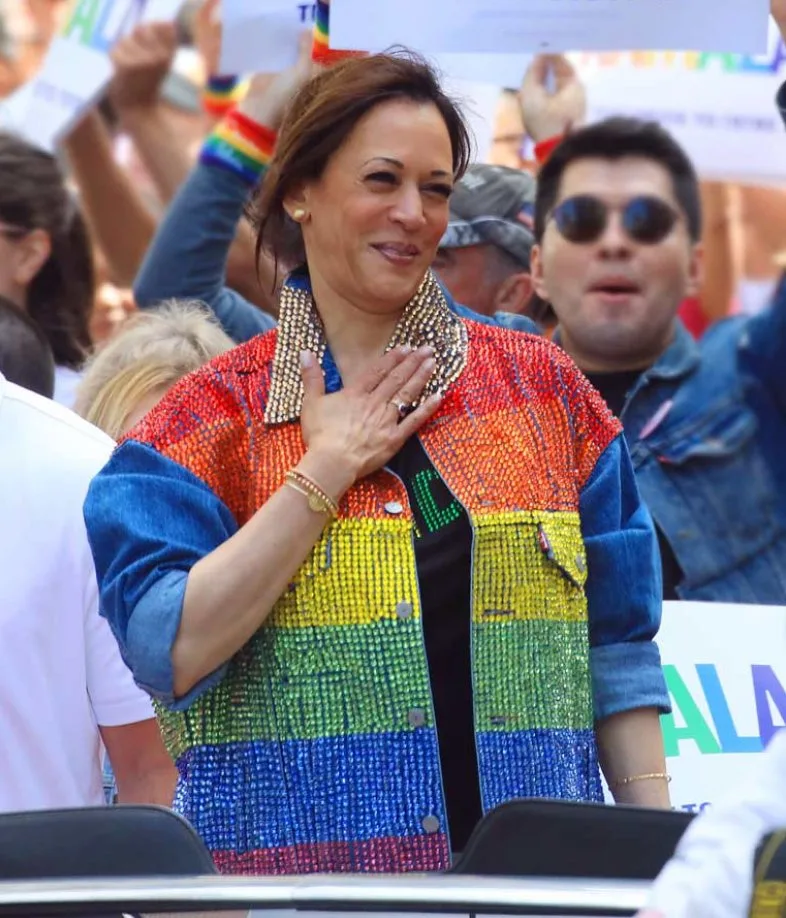 Kamala Harris smiling with a hand over her heart in a crowd at a Pride event, wearing a denim jacket whose front is covered in sequined horizontal stripes of red, orange, yellow, green, and blue, with rainbow-wristbanded supporters behind her.