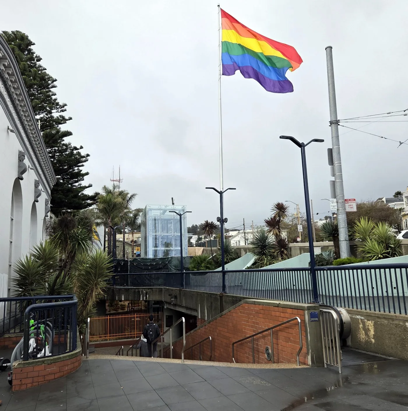 A giant six-stripe rainbow flag flying from a tall flagpole above Harvey Milk Plaza and the Castro Muni station entrance in San Francisco, under an overcast sky.