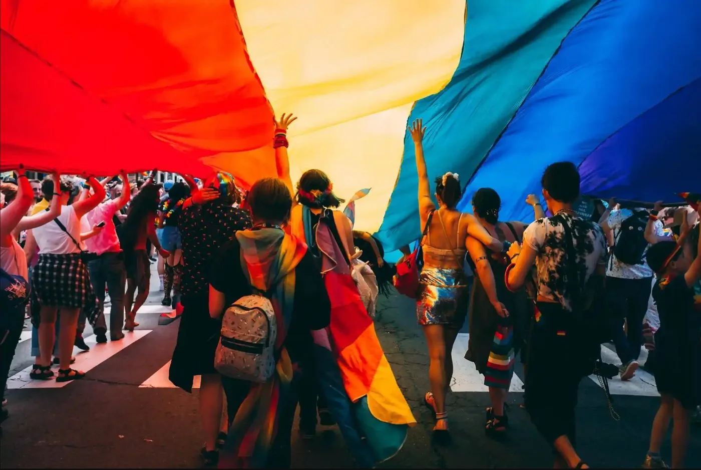 Marchers seen from beneath a giant rainbow flag they carry overhead at a pride parade, sunlight glowing through the red, yellow, and blue fabric.