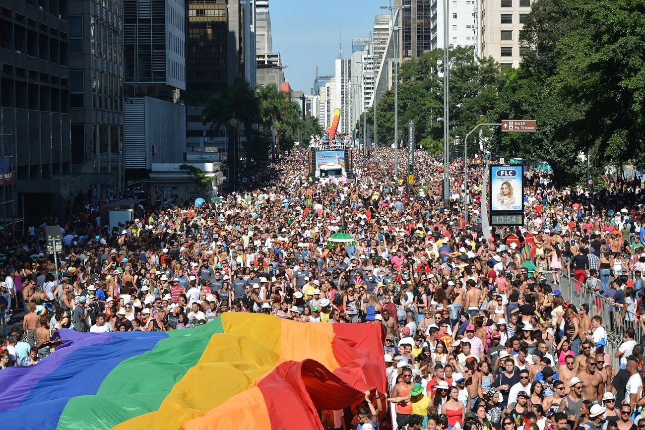 Looking down Avenida Paulista in São Paulo, packed shoulder to shoulder with a vast crowd, with an enormous rainbow flag being carried across the foreground.