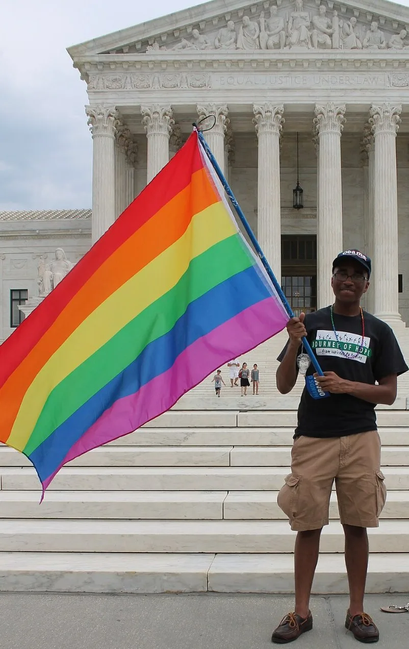 A young man in an Obama cap and a Journey of Hope t-shirt stands alone on the steps of the U.S. Supreme Court holding a large rainbow flag on a pole, beneath the pediment reading EQUAL JUSTICE UNDER LAW.