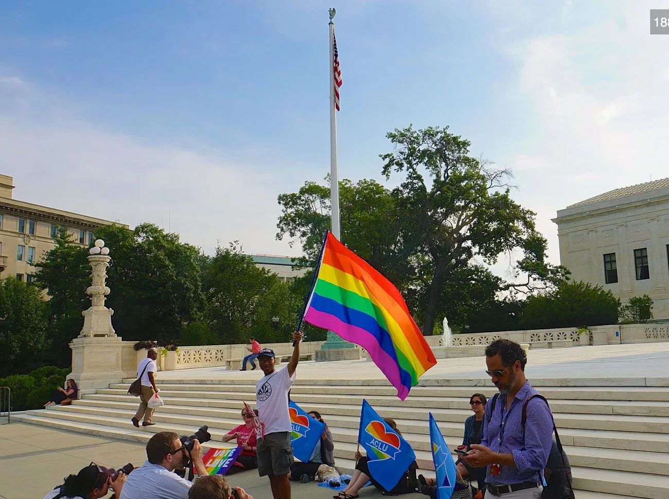 Wide morning view of the U.S. Supreme Court plaza: a man raises a rainbow flag beside the flagpole while press photographers kneel to shoot and supporters sit on the steps with blue ACLU flags.