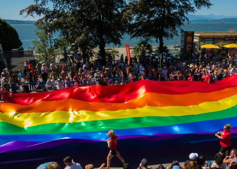A giant rainbow flag carried horizontally along a waterfront parade route in Vancouver, held at its edges by walkers, with spectators lining both sides and the sea behind.