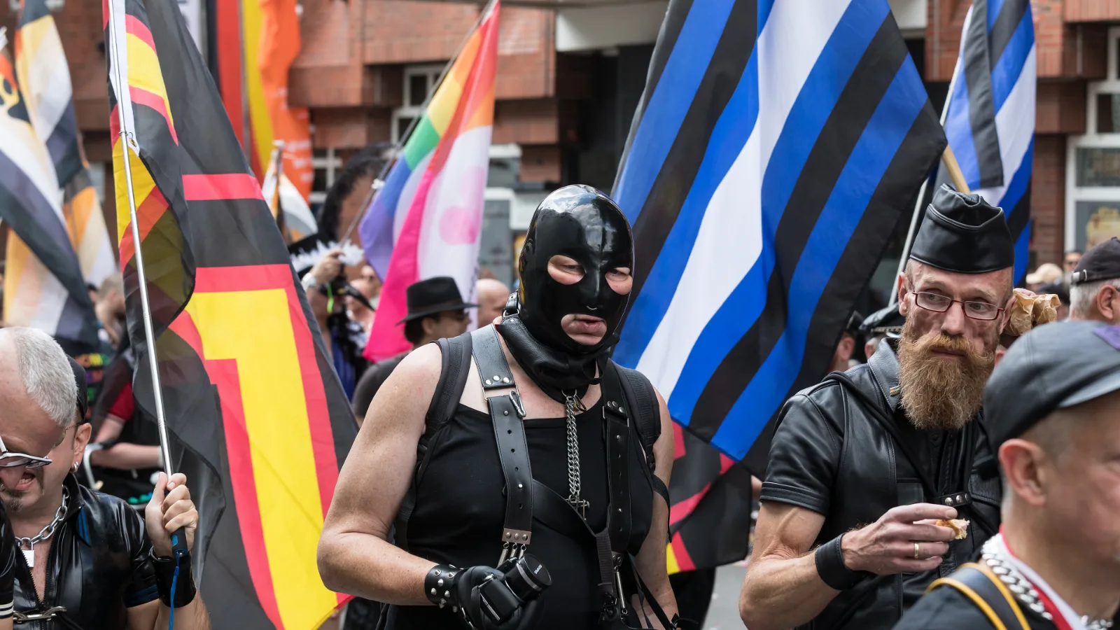 Marchers in leather and a black rubber hood at a Pride parade, with the rubber pride flag at left and the leather pride flag at right.