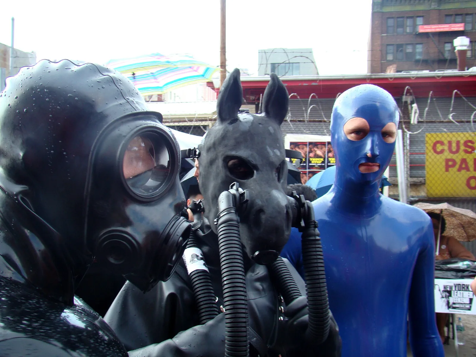 Three people in latex at a street fair: one in a black hood and gas mask, one in a horse mask with breathing tubes, one in a blue full-face latex hood.
