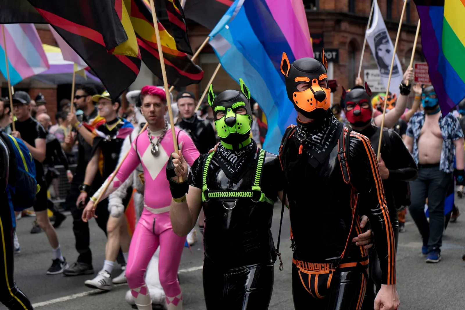 Marchers in latex and pup hoods at a Pride parade, with the rubber pride flag among transgender, non-binary and rainbow flags.