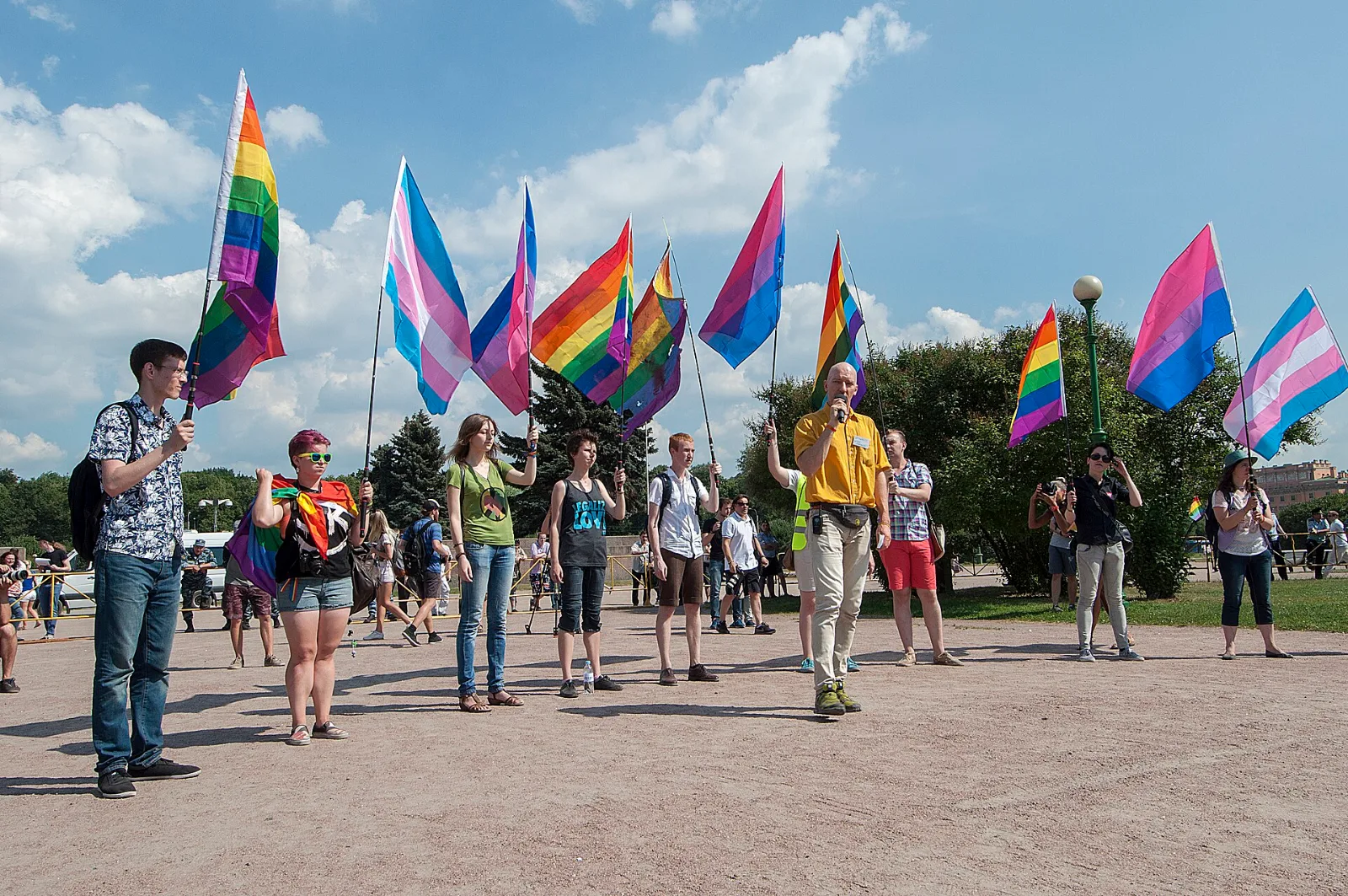 Roughly a dozen people standing in a line on open ground, each holding a flag on a pole: several rainbow flags, at least three transgender flags and several bisexual flags, against a bright sky.
