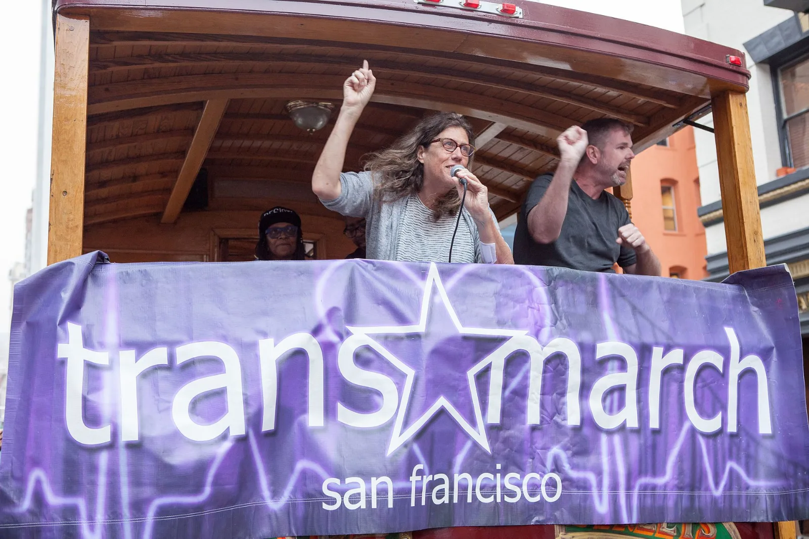 Susan Stryker speaks into a microphone with one arm raised, standing behind a purple banner reading trans march san francisco on the deck of an open cable car, with three other people beside her.