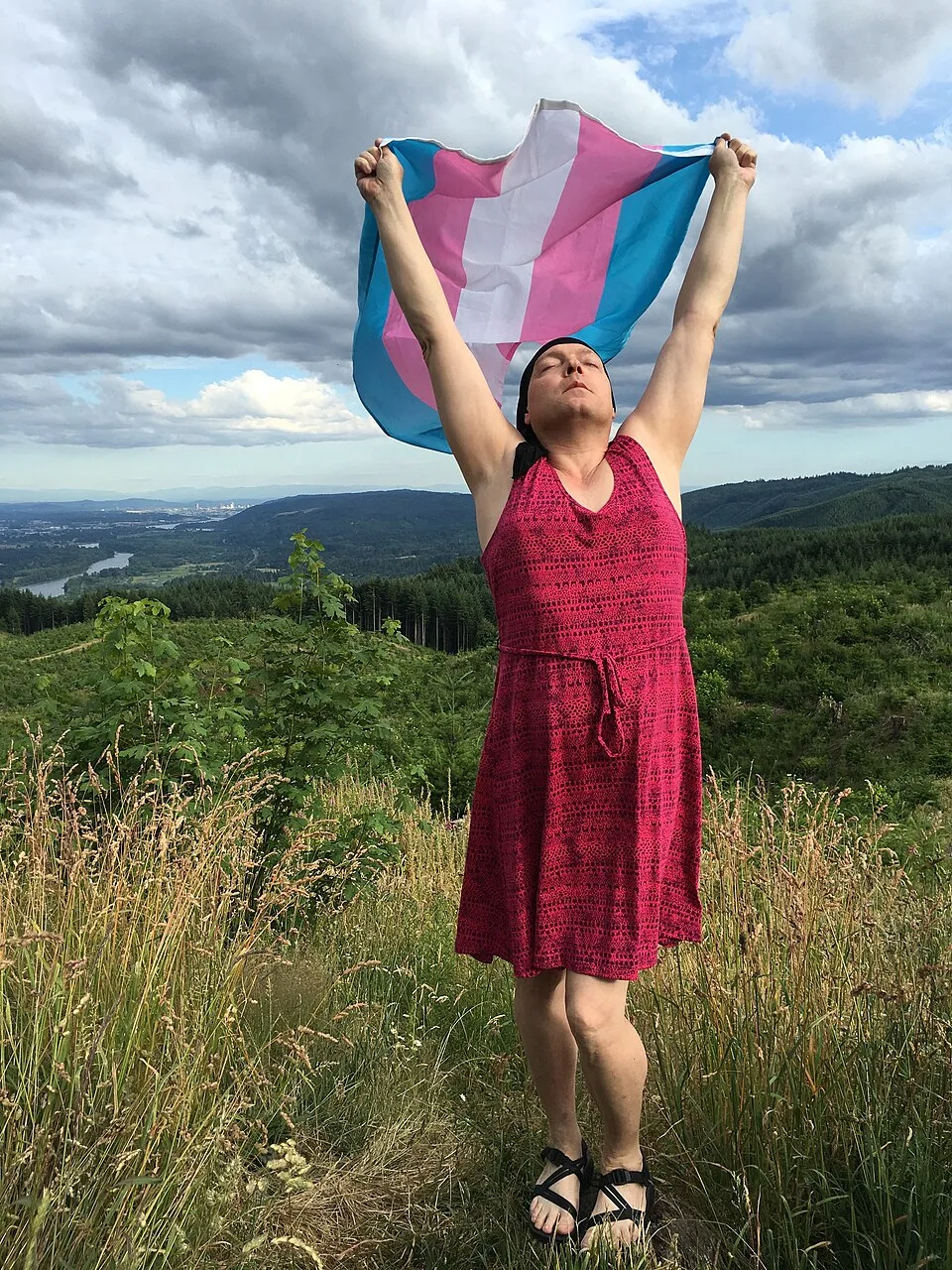 A person on a lawn holding a transgender pride flag spread open in both hands.