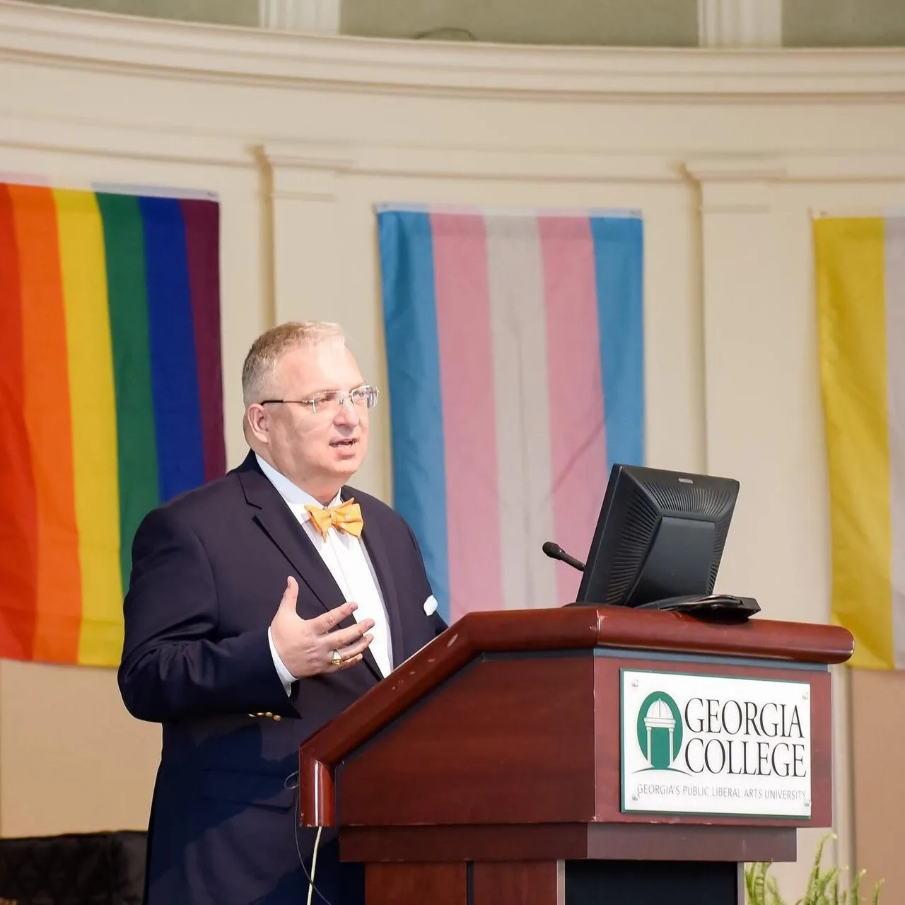 A speaker at a lectern in a ballroom, with pride and transgender flags displayed behind them.