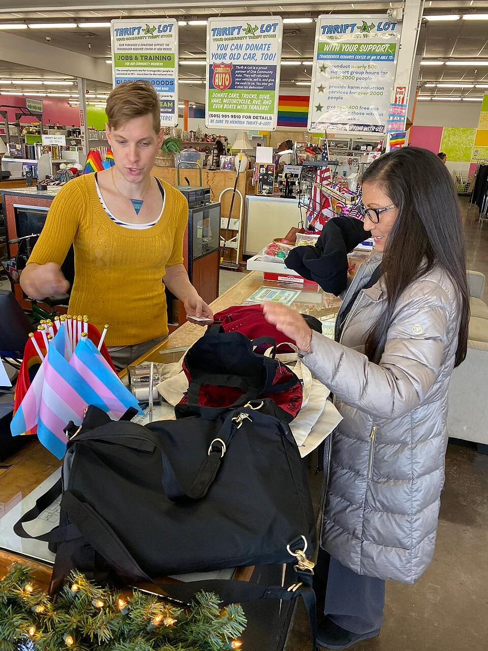 A person in a coat stands outside a shop holding a small transgender pride flag.