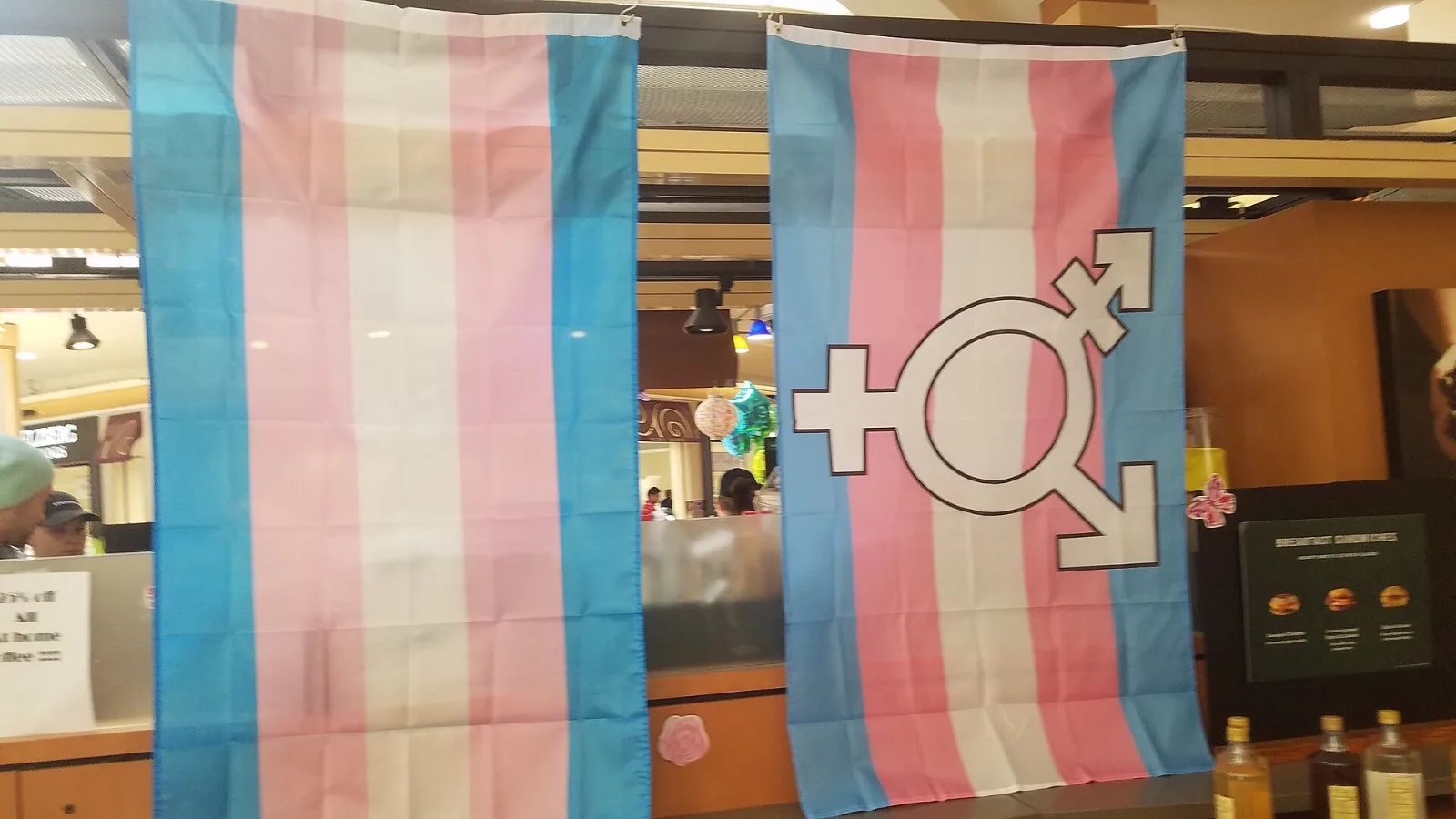 Two transgender pride flags flying outside a shop, one of them charged with the transgender symbol.