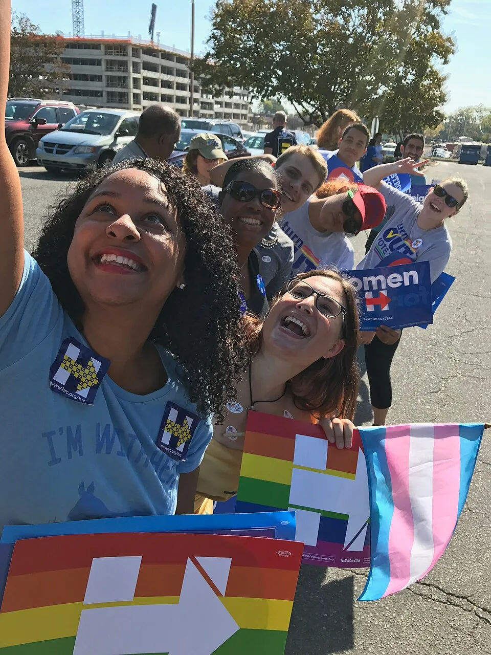 A person holding a transgender pride flag at an outdoor campaign event.