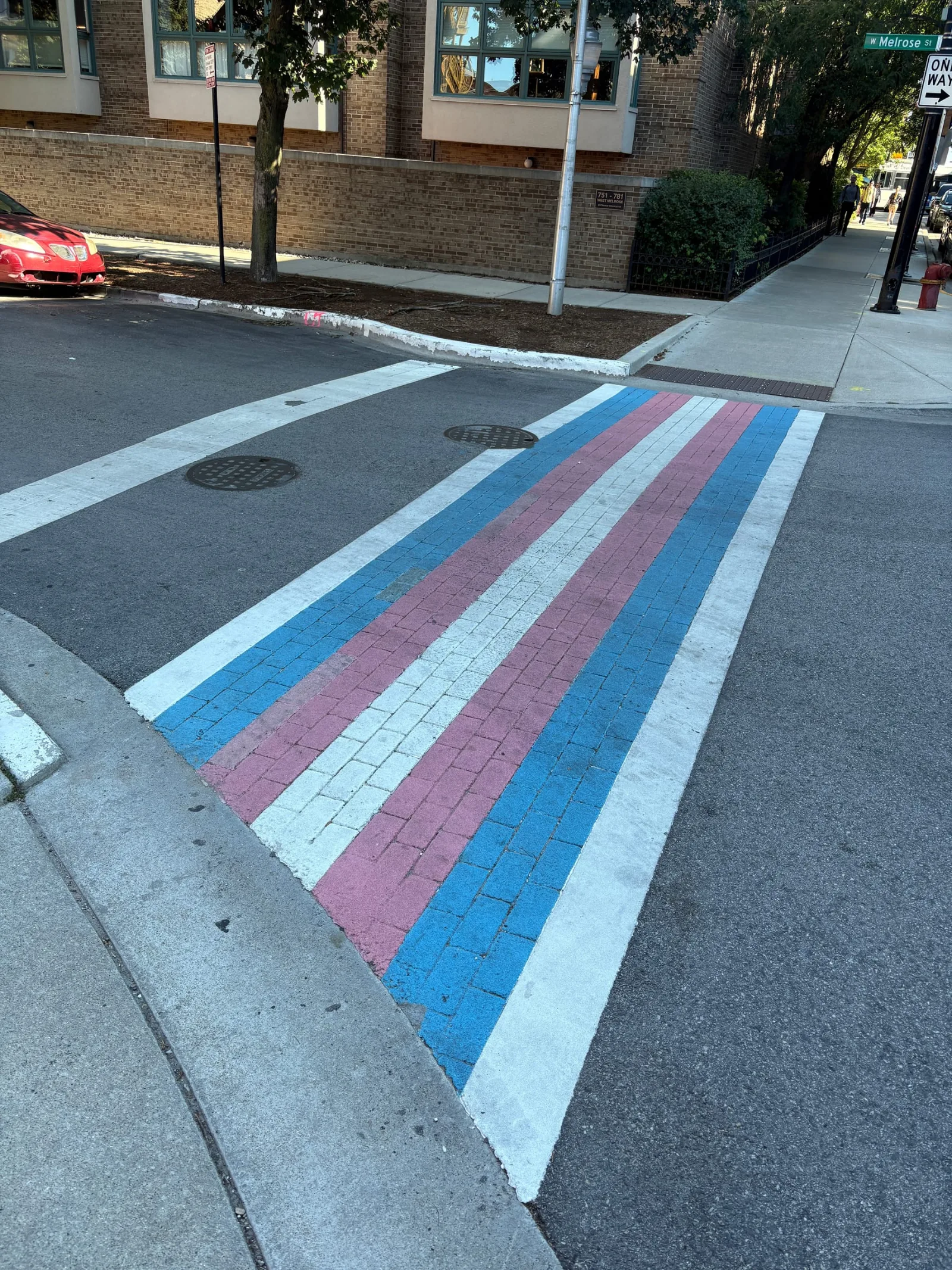 A transgender pride crosswalk laid into brick paving at North Halsted Street and West Melrose Street in Chicago, in light blue, pink and white stripes, with the West Melrose street sign on the far corner.