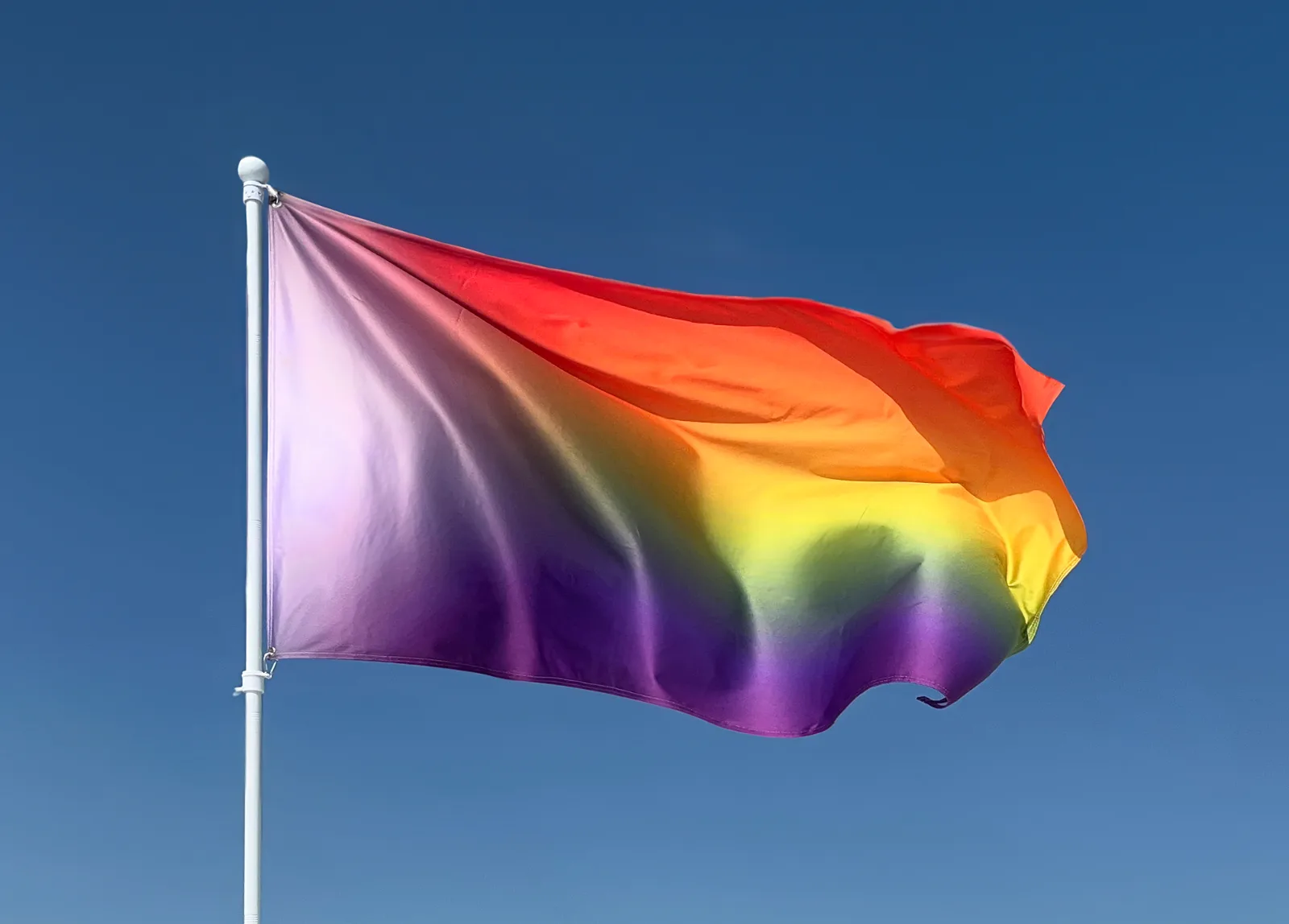 Utopian Pride Flag #6 flying from a white flagpole against a clear blue sky, its blurred rainbow running red at the top through yellow to purple, with the pale chevron region at the hoist.