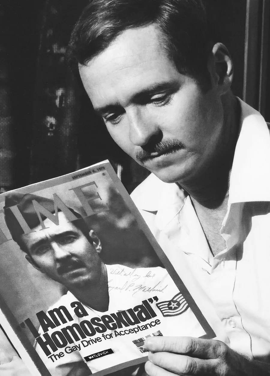 Black-and-white photograph of Leonard Matlovich in an open-collared white shirt, looking down at an autographed copy of his 1975 Time cover held in his hand.