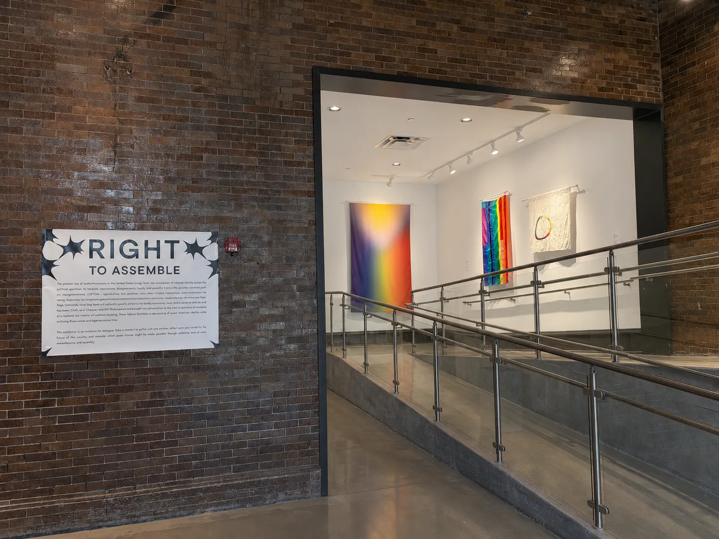 Right to Assemble installation view: the gallery entrance with the exhibition title on a brick wall, the 5790/2029 Pride Flag and Utopian Pride Flag #25 hanging beyond.