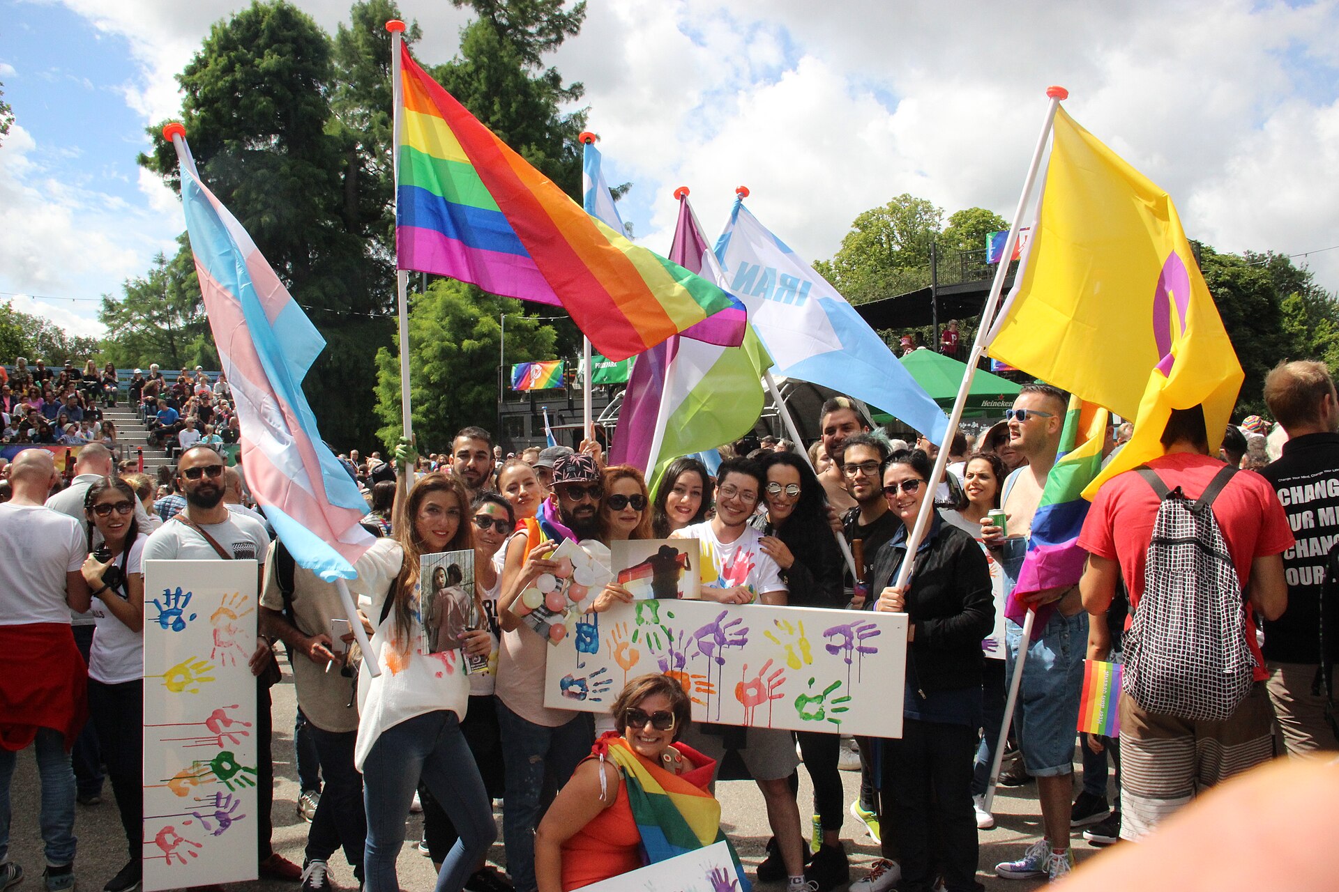 A yellow flag with a purple ring carried among Pride flags in a park.