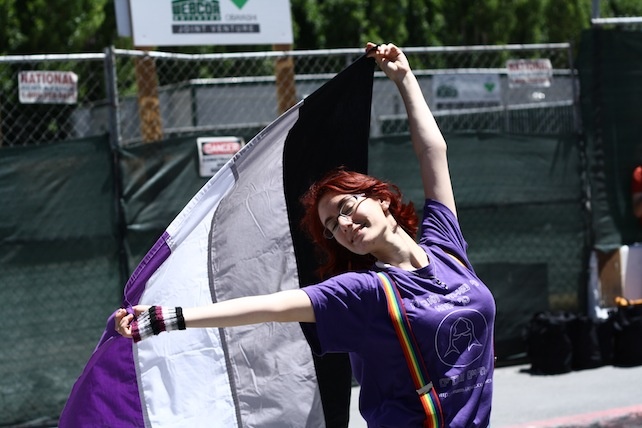 A person in a purple shirt with rainbow suspenders joyously waving a large asexual flag at an outdoor event.