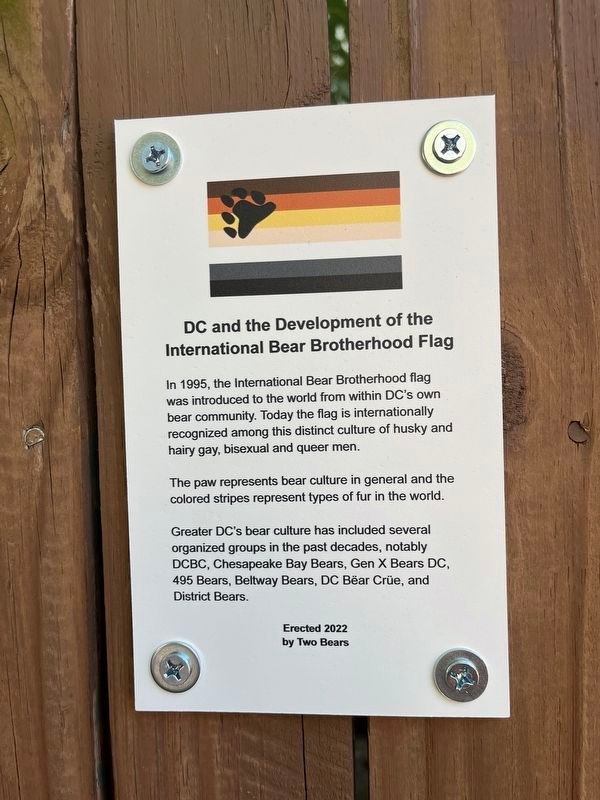 A white historical marker mounted on a wooden fence, headed "DC and the Development of the International Bear Brotherhood Flag," with the flag reproduced above the text.