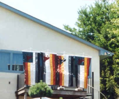 Four striped flags with paw prints hung vertically from a line under the eaves of a house, above a wooden deck.