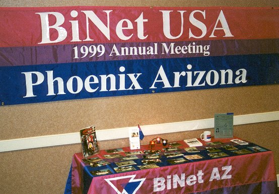 A banner in magenta, purple and blue reading BiNet USA 1999 Annual Meeting, Phoenix Arizona, above a table draped in the same colors.