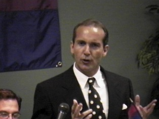 Michael Page in a dark suit speaking into a microphone, with a bisexual pride flag hanging on the wall behind him.