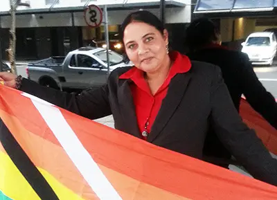 A woman in a black jacket and red shirt standing on a city street holding the flag open in front of her.