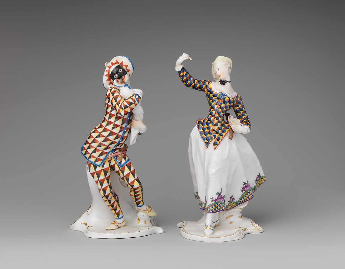 Two porcelain figures side by side. At the left a masked male harlequin in a hat and a suit patterned throughout in red, yellow and black triangles, carrying a white bundle; at the right the Harlequina in her blue and yellow triangle jacket and white skirt.
