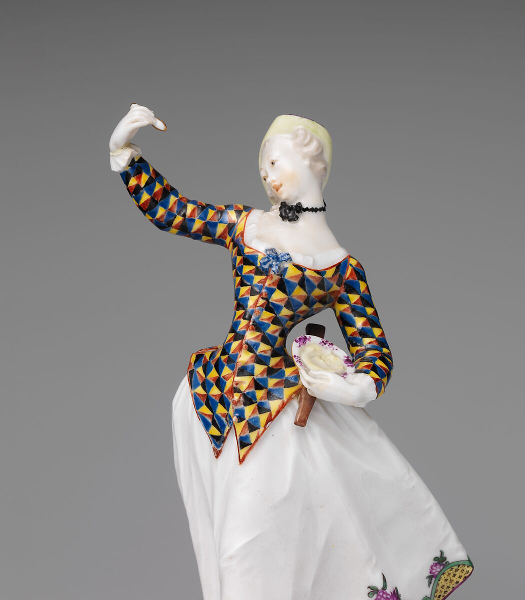 Detail of a porcelain figure: a woman's fitted jacket and sleeves painted in a dense tessellation of triangles in blue, yellow, black and red, worn over a white skirt, with a black choker at the throat and one gloved hand raised.