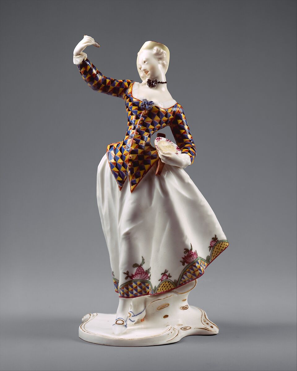 A porcelain figure of a woman in a triangle-patterned jacket of blue, yellow and dark blue over a full white skirt bordered with flowers, one arm raised and a slapstick and dish held at her waist, standing on a scrolled white base.