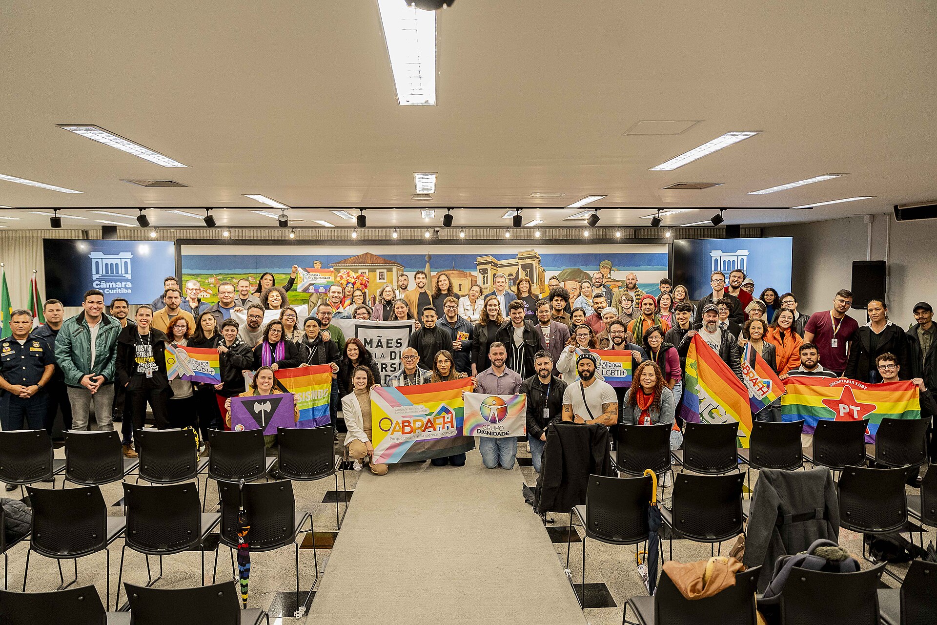 A large group photograph in a city council chamber, rows of black seats in the foreground. Toward the left of the group two people hold a violet flag with a black inverted triangle and white double-headed axe. Around it are rainbow banners lettered ABRAFH, Grupo Dignidade, Aliança LGBTI+ and Secretaria LGBT PT, and a black banner reading MÃES DA DIVERSIDADE.