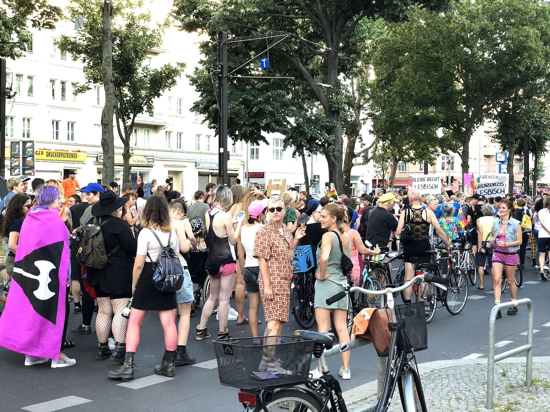 A person at the head of a crowded street march wears a magenta flag as a cape, its black inverted triangle and white double-headed axe turned toward the camera. Marchers with bicycles fill the road behind; hand-lettered placards at the right read BLEIBE RECHT LESBISCH and GRENZENLOS LESBISCH.