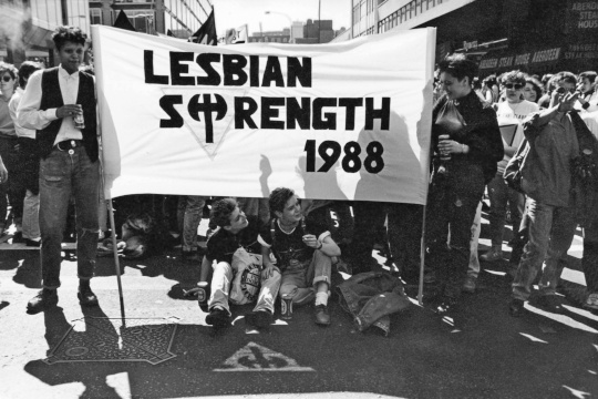 A black and white photograph of a street march halted. Two people hold a large white banner lettered LESBIAN STRENGTH 1988, in which the T of STRENGTH is drawn as a double-headed axe standing on an inverted triangle. Two people sit on the road in front of the banner; a third inverted triangle is marked on the road surface in the foreground. Marchers and shopfronts fill the background.