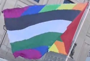 A photograph of the flag flown outdoors: a rainbow field with slanted stripes, carrying the Palestinian flag's black, white and green bands and a red chevron.