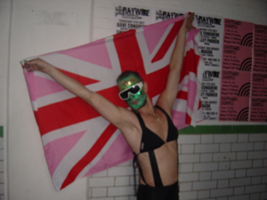 A performer in green face paint holding a Pink Jack outstretched against a poster wall.