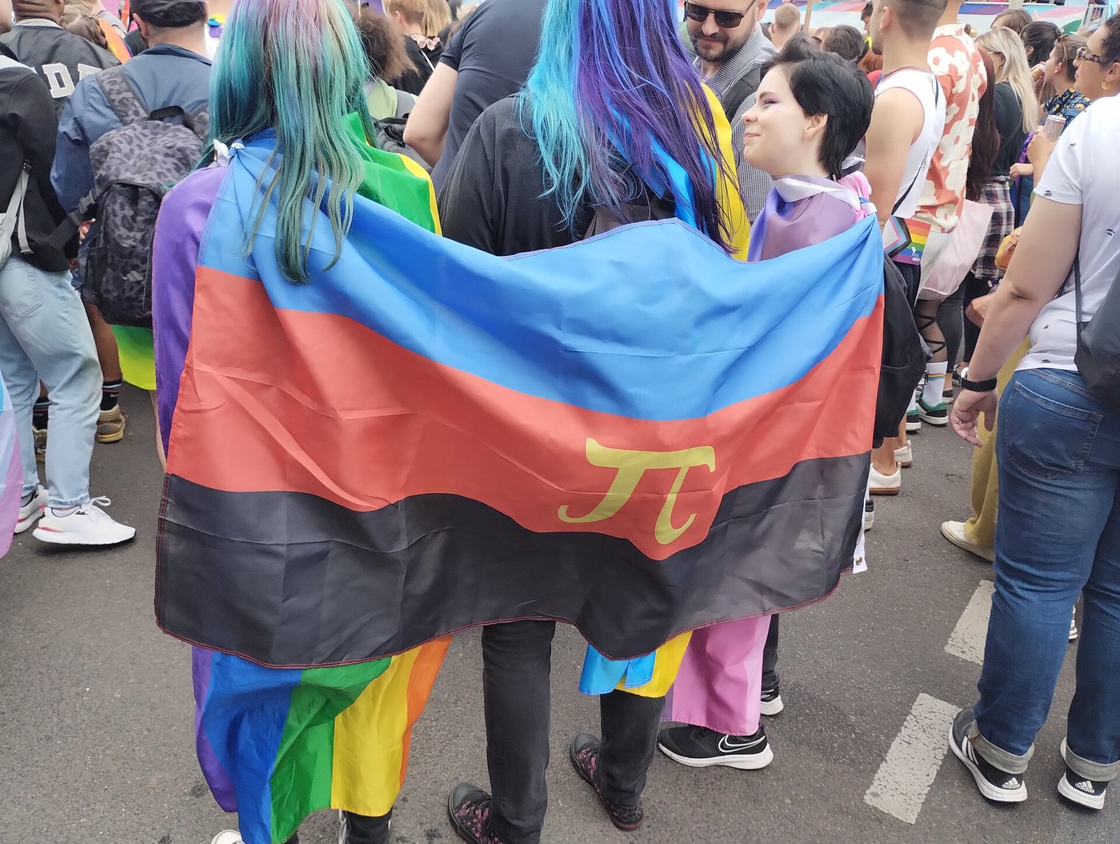 Three people at a Pride parade wearing a large blue, red and black flag with a gold Greek pi as a cape.