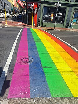 A pedestrian crossing painted in six rainbow stripes across a street corner, with parked cars and low buildings behind.