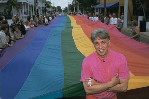 Gilbert Baker in a pink shirt, arms crossed, standing in front of an enormous rainbow flag that stretches away down a crowded street held at waist height by onlookers on both sides.